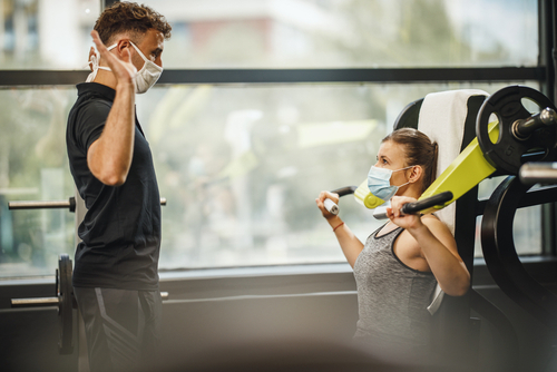 Shot of a muscular young woman with protective mask working out with personal trainer at the gym machine during Covid-19 pandemic. She is pumping up her shoulder muscule with heavy weight.