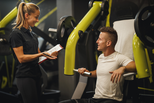 Shot of a muscular young woman in sportswear working out with personal trainer at the gym machine. She is pumping up her chest muscule with heavy weight.