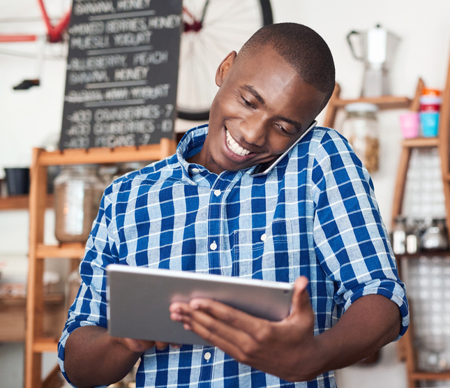 Smiling young African entrepreneur talking on a cellphone and using a digital tablet while standing at the counter of his cafe