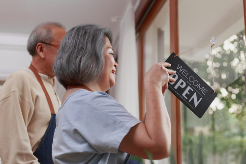 Happy Asian retired senior Asian couple standing in the house as a small business restaurant putting opening sign on the door with happy welcoming smiley face.