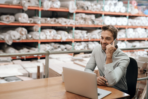 Smiling manager working at his desk in a carpet warehouse and talking on a cellphone with shelves of stock in the background